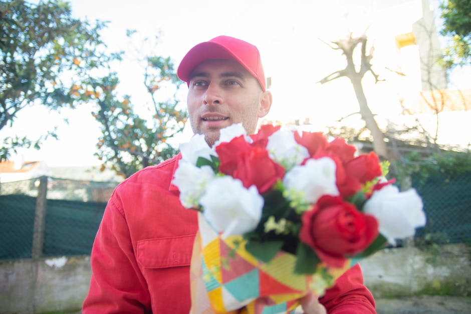 A delivery person dressed in a red jacket and matching red cap is holding a vibrant floral bouquet with a mix of red and white roses, elegantly arranged in a colorful, patterned wrapping paper. The bo