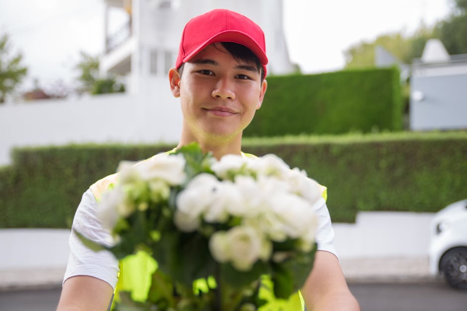 A cheerful delivery person wearing a red cap and a yellow high-visibility vest holds out a bouquet of fresh white roses, green foliage, and other white flowers. The bouquet is wrapped in clear and whi