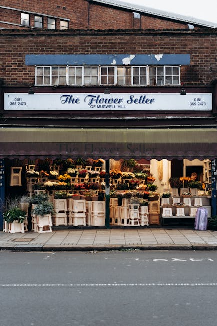 The storefront of The Flower Seller of Muswell Hill, featuring a variety of fresh floral arrangements displayed on white wooden stands and shelves. The flowers include vibrant yellow roses, purple lav