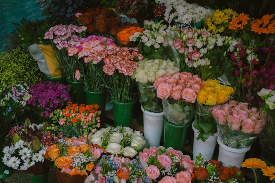 A diverse arrangement of fresh flowers displayed on a floral shop shelf, featuring bouquets of pink, white, yellow, orange, and purple blooms. The flowers include roses, carnations, daisies, and other