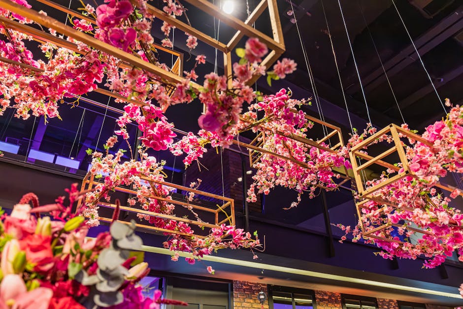 An interior ceiling decorated with hanging arrangements of pink cherry blossom branches, suspended inside geometrically framed wooden structures. The vibrant pink flowers contrast with the dark ceilin