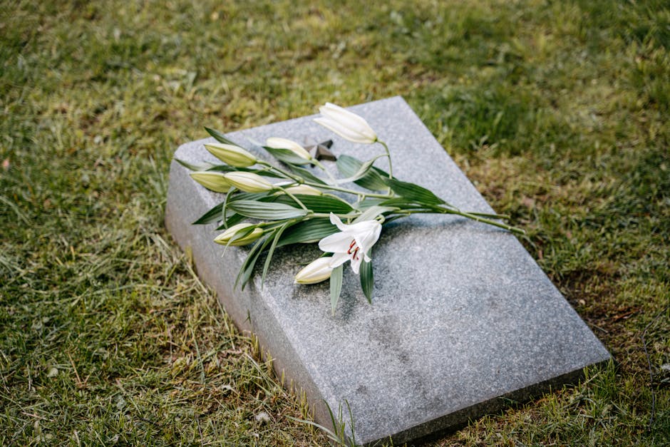 A bouquet of fresh white lilies with long green leaves and closed buds, resting on a flat, gray granite tombstone in a grassy area. The lilies have open petals revealing dark stamens, and the arrangem
