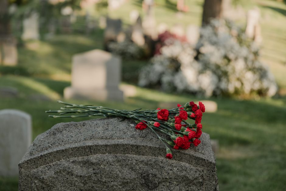 A close-up view of a bouquet of fresh red carnations lying on a gray stone grave marker at a cemetery. The carnations have vibrant, slightly ruffled petals with green stems and leaves, arranged casual