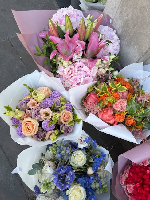 A display of five beautifully arranged floral bouquets on a concrete surface, featuring a variety of fresh flowers. The central bouquet prominently showcases pink lilies with large, open petals, surro