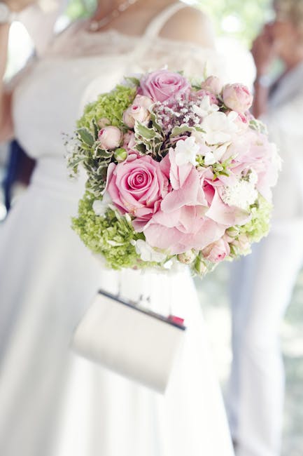 A floral bouquet arrangement featuring large, soft pink roses, pale pink hydrangeas, and smaller white and pink buds, accented with green foliage and baby's breath. The bouquet is held by a person dre