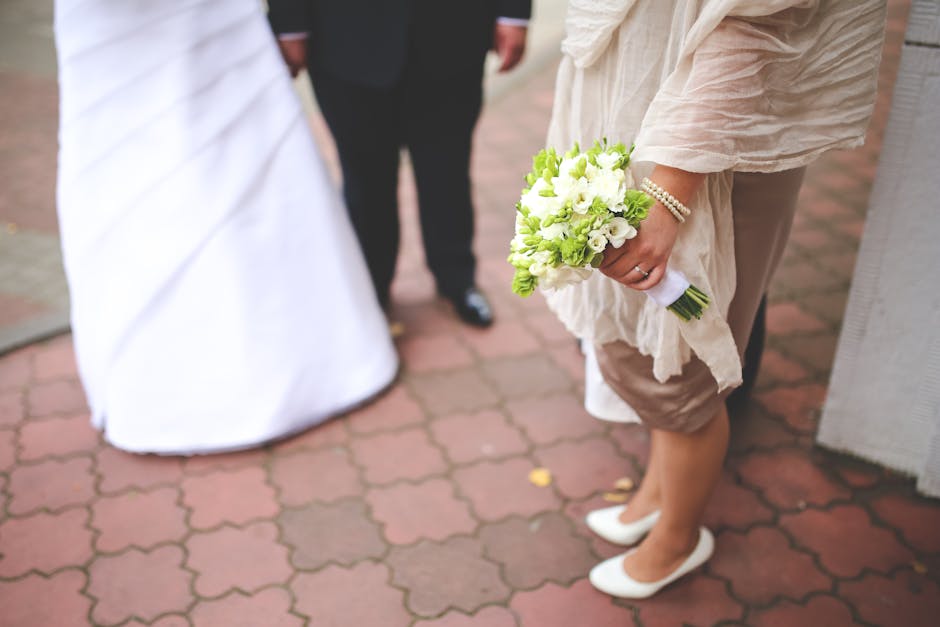 A person holding a bouquet of fresh white and light green flowers, including roses and freesias, wrapped in white paper and secured with a green ribbon, is standing on a red brick pavement during a we