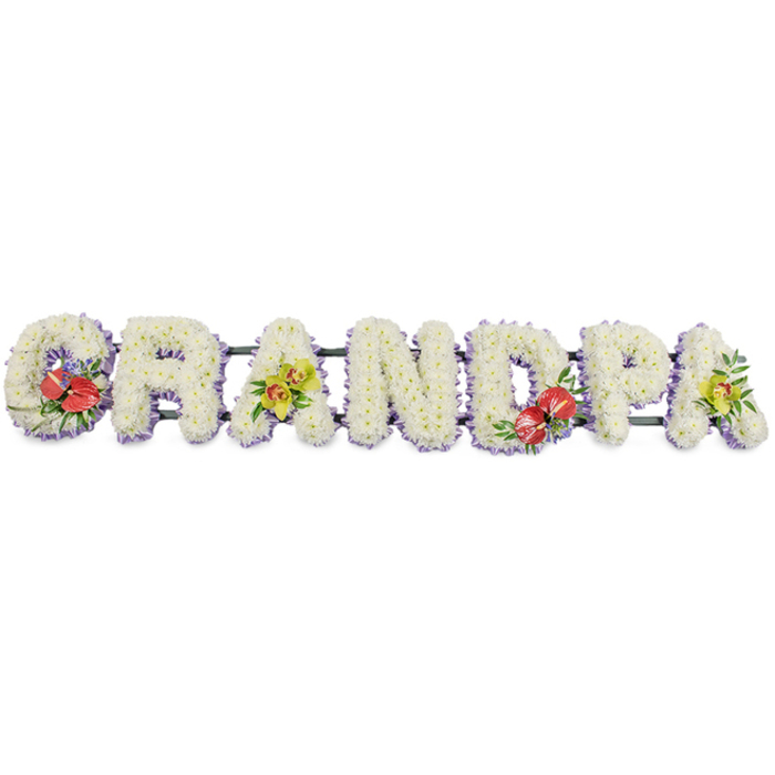 A personalised sympathy tribute spelling out GRANDPA stretches horizontally across the frame, each letter formed as a deep, blocky structure overflowing with fresh white blooms. The letters stand shoulder to shoulder in a continuous line, aligned on hidden dark green supports that lend strength while keeping the visual focus on the flowers. Every capital is packed tightly with small white chrysanthemums, their neatly layered petals creating a velvety, almost quilted finish. The consistent use of this single flower type gives the word a sense of unity and serenity, as if the entire tribute were a single soft cushion of white. Along the base of each letter, a frill of pale lavender ribbon curves gently around the form, just visible beneath the petals. This subtle purple edging adds light contrast and outlines the word with a calm, cool tone that softens the edges without disturbing the simplicity of the design. The overall palette is quiet and refined, but four concentrated accent sprays introduce warmth and personality. On the 'G' and 'D', clusters of deep red anthuriums with their glossy, heart-shaped spathes and golden-yellow spadices emerge dramatically from the white background, surrounded by lush green foliage and a few delicate filler flowers. On the first and final 'A', elegant yellow-green orchids appear, their slender petals and speckled throats curving upward, supported by fresh greenery that spills gently over the edge of the letters. This style of floral lettering is a familiar sight at funerals and memorial services across Kennington, from intimate gatherings near Cleaver Square to larger services along Kennington Park Road, created to honour a beloved grandfather with a message of love, respect, and lasting remembrance written in flowers.